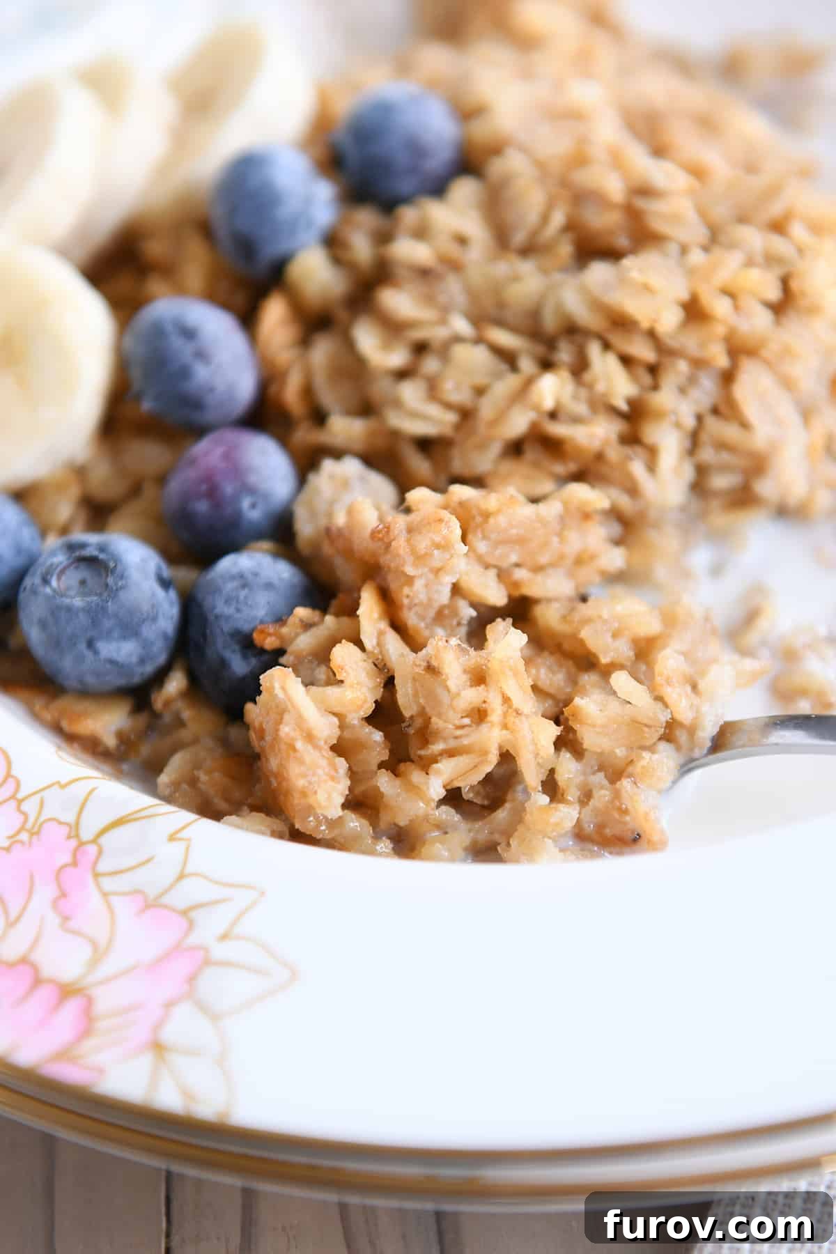 A spoon laden with baked oatmeal, fresh blueberries, and sliced bananas, poised above a beautifully patterned floral bowl.