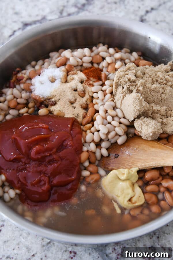 A shallow pan filled with the unbaked ingredients for saucy baked beans, including white beans, brown sugar, ketchup, and Dijon mustard, awaiting baking. A wooden spoon rests in the mixture.