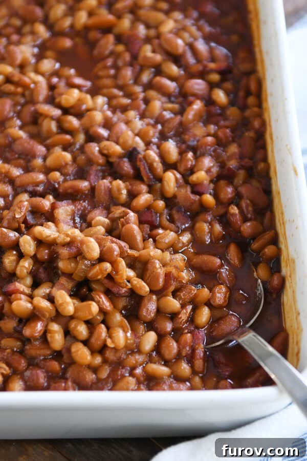 A serving spoon nestled in the corner of a white pan filled with steaming, saucy baked beans, freshly out of the oven.
