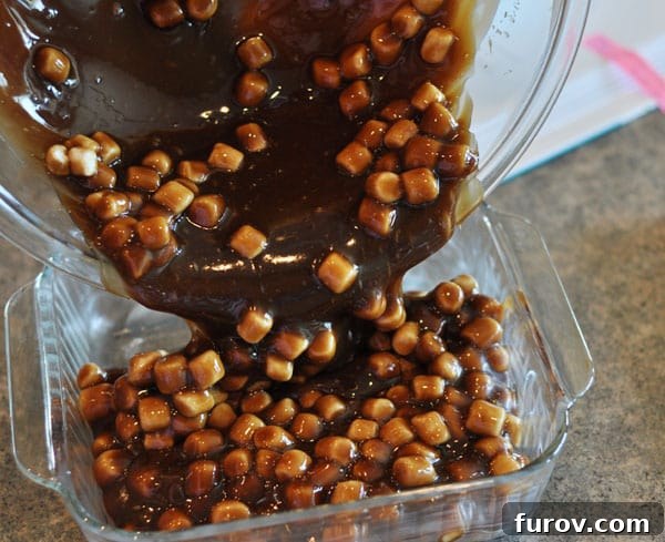 Marshmallow studded fudge being smoothly poured into a glass 8x8 inch pan, spreading to fill the bottom.