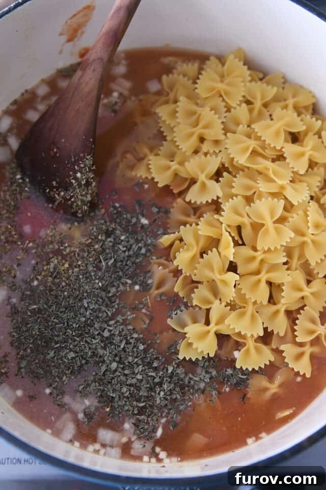 Bowtie pasta, chicken broth, crushed tomatoes, oregano, and basil simmering in a cast iron pot.