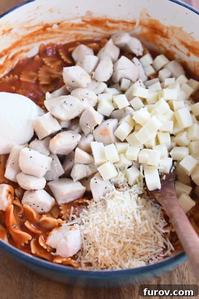 Cubed mozzarella, cooked chicken, parmesan cheese, and ricotta cheese being stirred into bowtie pasta in a cast iron pot.