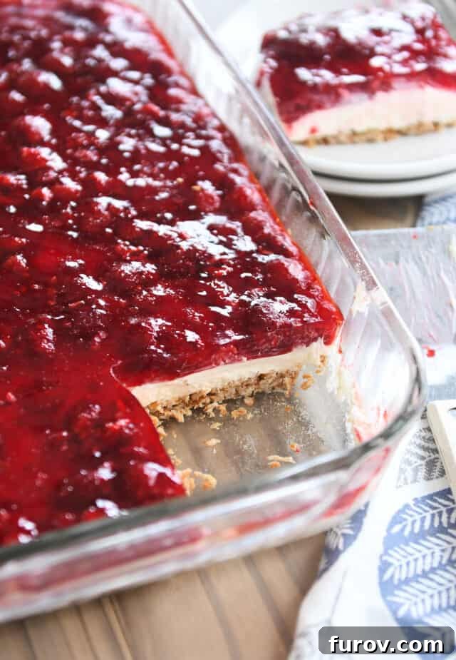 A 9x13-inch pan filled with vibrant Raspberry Pretzel Jello, a slice removed to reveal its distinct layers: a golden-brown pretzel crust, a creamy white filling, and a ruby-red raspberry jello topping.