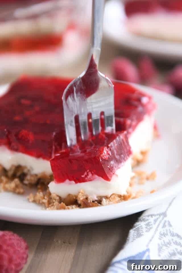 A fork gently scooping a portion from a square of Raspberry Pretzel Jello on a white plate, highlighting the layers and inviting a taste.