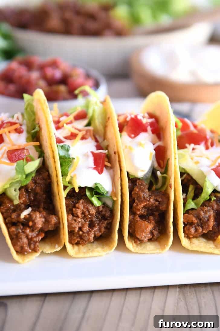 Four crispy-shelled ground beef tacos filled with fresh toppings on a pristine white platter, showcasing the rich, perfectly seasoned meat.