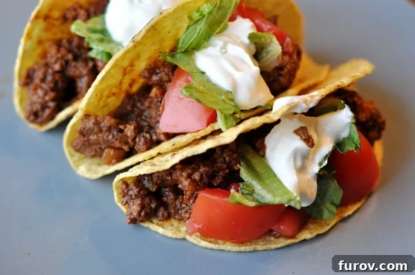 Three ground beef tacos generously topped with cool sour cream, crisp lettuce, and diced fresh tomatoes, presented on a rustic wooden board.