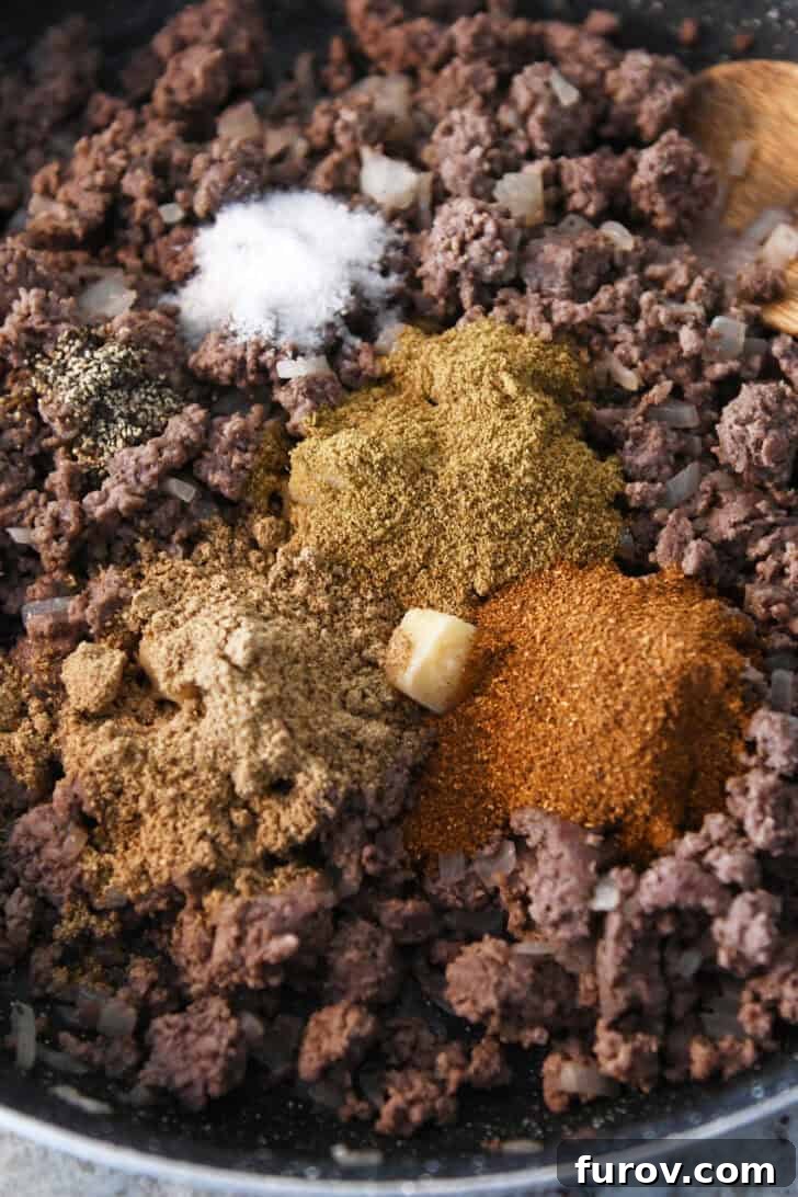 Top-down view of perfectly cooked ground beef with a vibrant mix of seasonings being poured into the center, ready to be stirred in and create incredible taco meat.
