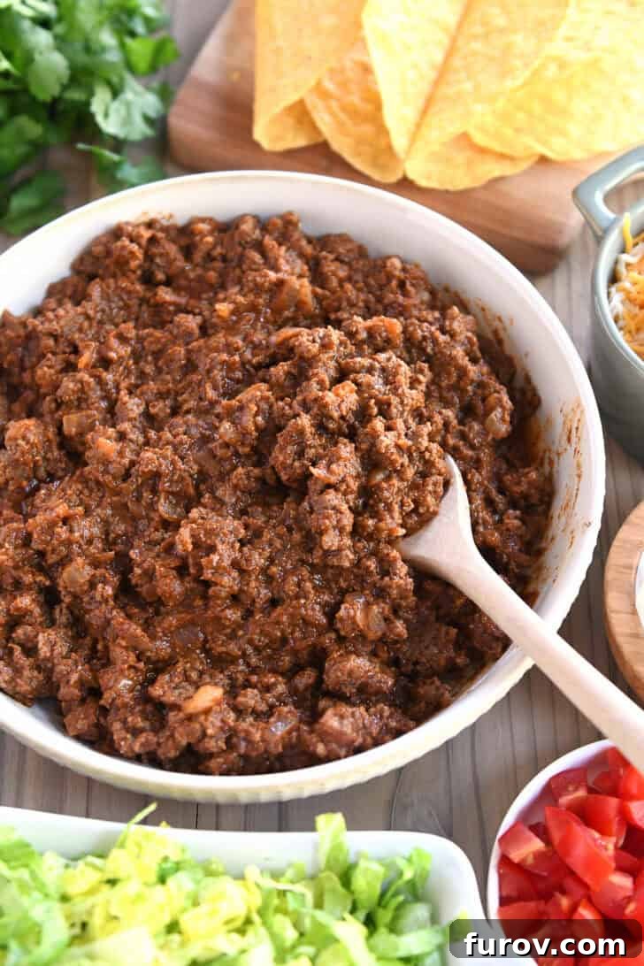 Top-down view of a pristine white bowl generously filled with seasoned ground taco meat, surrounded by an array of colorful taco toppings in smaller bowls, creating a vibrant taco bar spread.