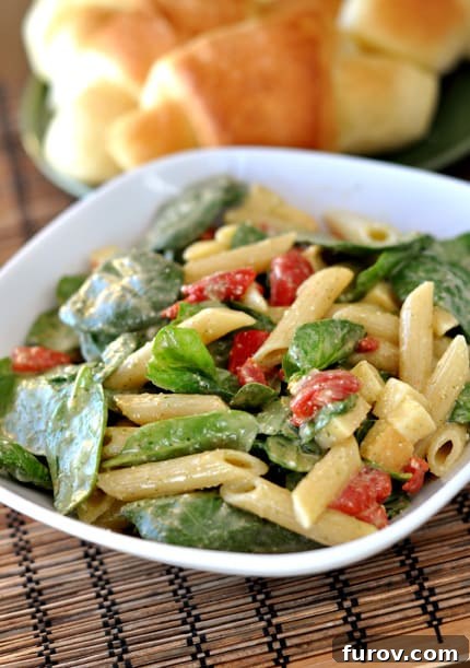 A beautifully arranged white dish showcasing a vibrant penne, spinach, and roasted red pepper salad, served with a side of golden crescent rolls, ready to be enjoyed.