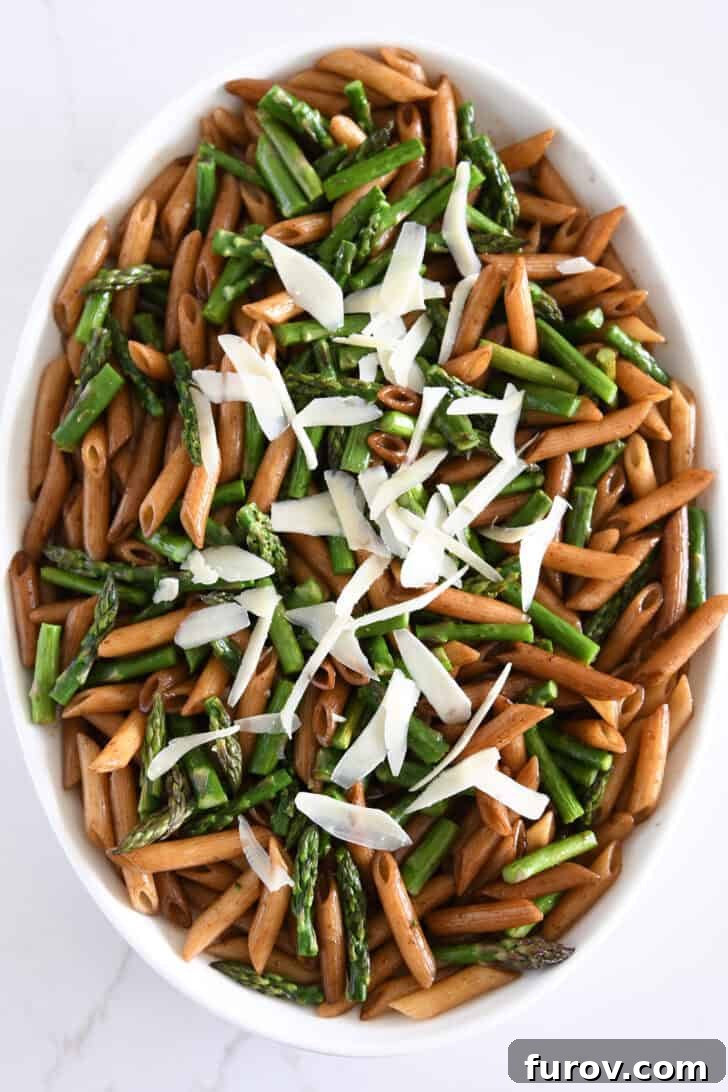 Top-down view of penne pasta in a white serving dish with roasted asparagus, balsamic butter sauce, and freshly grated Parmesan cheese.
