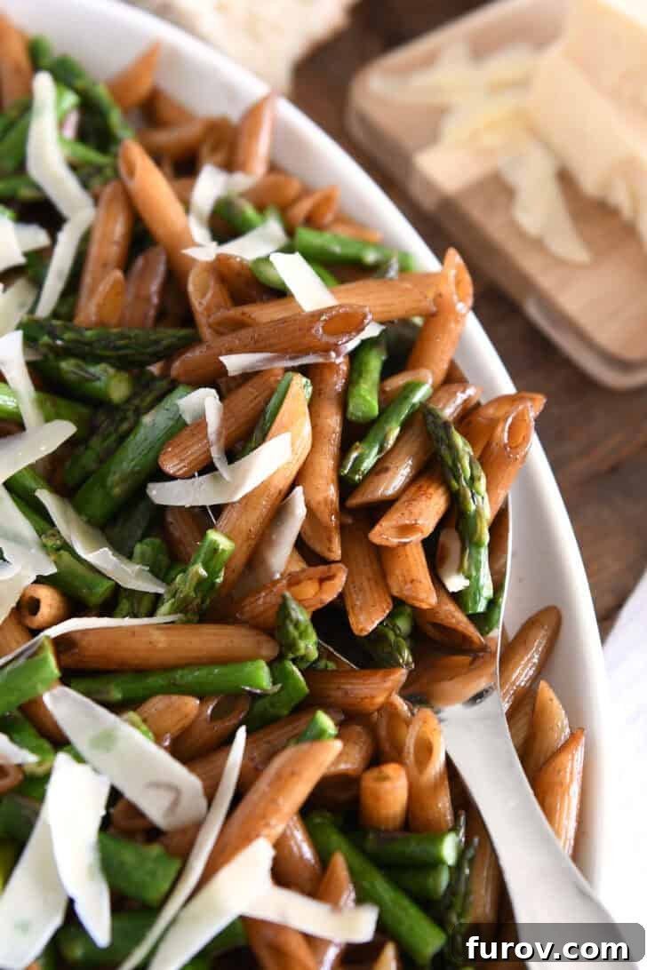 Serving spoon lifting a portion of balsamic penne pasta, roasted asparagus, and parmesan cheese from a white dish.