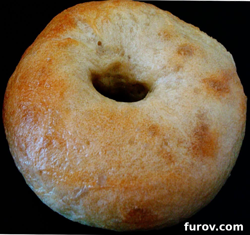 A single perfectly golden-brown homemade bagel with a dark, chewy crust rests on a black plate, showcasing its appealing texture and classic hole.