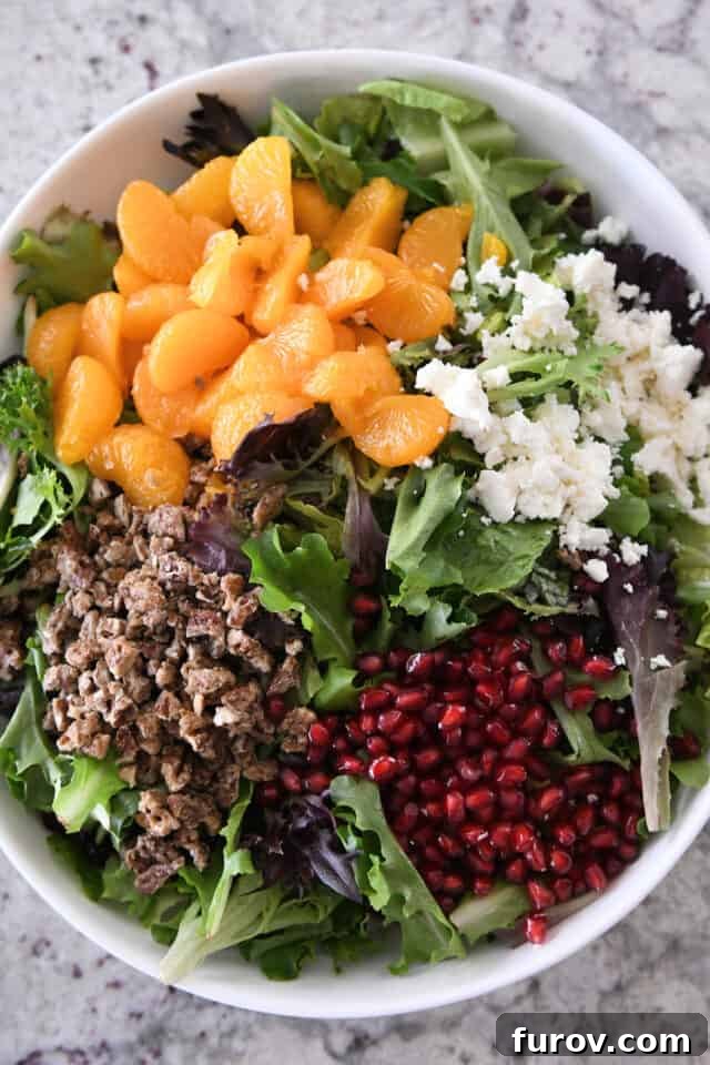 A large white salad bowl overflowing with a vibrant holiday salad: mixed greens, mandarin oranges, crumbled feta cheese, candied pecans, and glistening pomegranate arils, ready to be served.