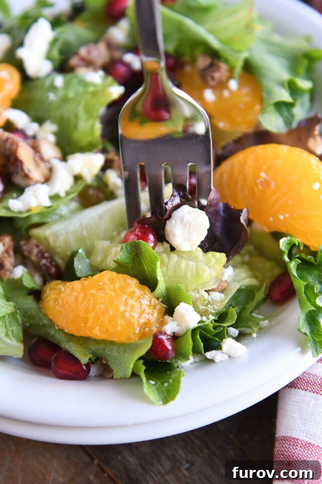 A fork scooping a perfect bite of holiday salad, featuring mixed greens, a piece of crumbled feta cheese, a juicy mandarin orange segment, and glistening pomegranate seeds.