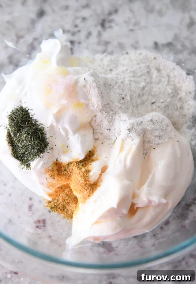 A pristine glass bowl filled with the creamy, white mixture of mayonnaise and sour cream, surrounded by small piles of dry ingredients: bright green dill, savory seasoning salt, and the aromatic white powder of ranch seasoning.