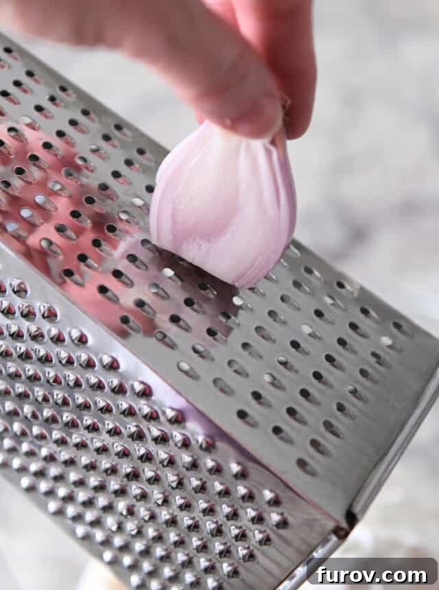 A close-up view of a hand expertly grating a small, golden-brown shallot against the fine holes of a stainless steel box grater, preparing the aromatic ingredient for the creamy veggie dip.