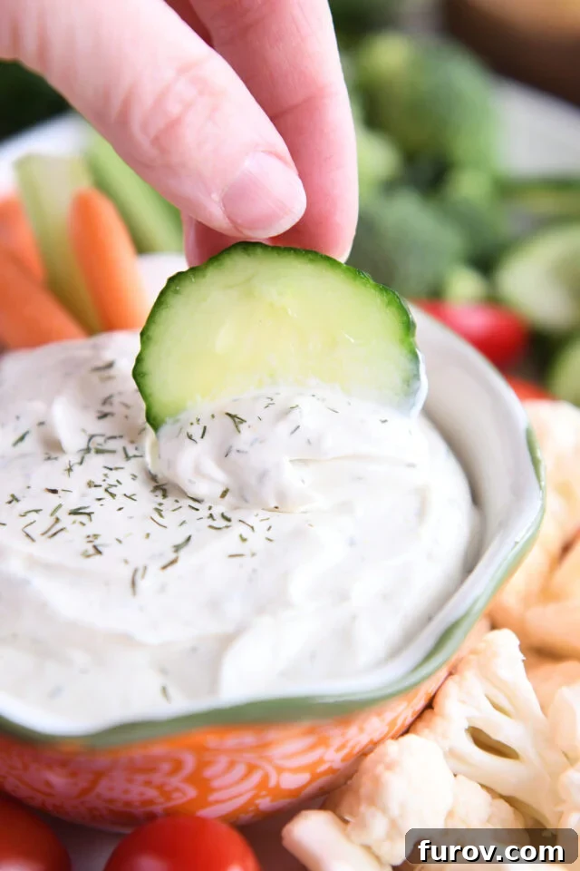 A vibrant green cucumber slice being dipped into a rich, creamy veggie dip presented in a striking orange bowl, set against a clean background.