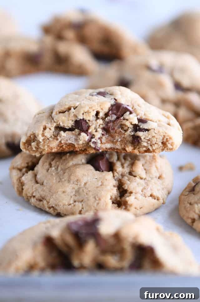 A perfectly baked vegan peanut butter oatmeal chocolate chip cookie, broken in half, resting on a whole cookie, on a parchment-lined baking sheet, showcasing its soft interior and gooey chocolate chips.