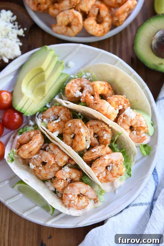 Three perfectly assembled shrimp tacos on a pristine white plate, garnished with fresh avocado slices and juicy cherry tomatoes, highlighting the vibrant colors and textures of the dish.