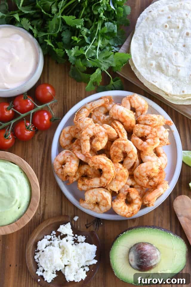 A vibrant plate artfully arranged with cooked shrimp, bright cherry tomatoes, a bowl of creamy avocado sauce, warm tortillas, fresh cilantro, and crumbled cotija cheese, ready for building the ultimate shrimp tacos.