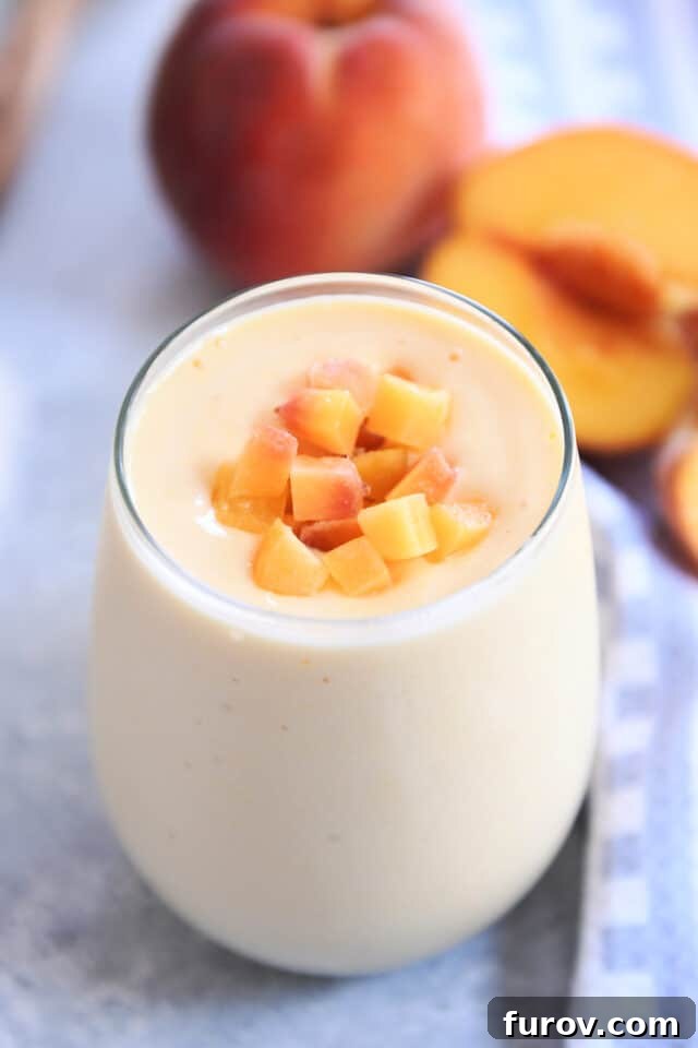 A vibrant peach orange protein smoothie in a clear glass cup, garnished with fresh diced peaches. Peaches are artfully arranged in the soft-focus background.