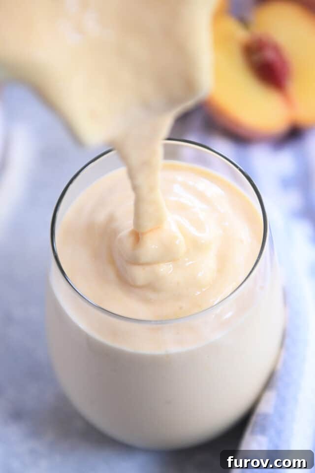 Someone is carefully pouring the freshly blended, smooth peach orange protein smoothie into a tall, clear glass cup.