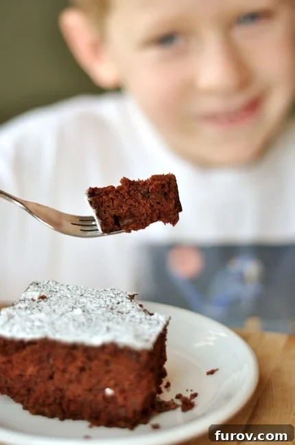 A piece of powdered sugar dusted chocolate cake with a little boy behind it holding a bite of the cake on his fork, proudly showing his creation.
