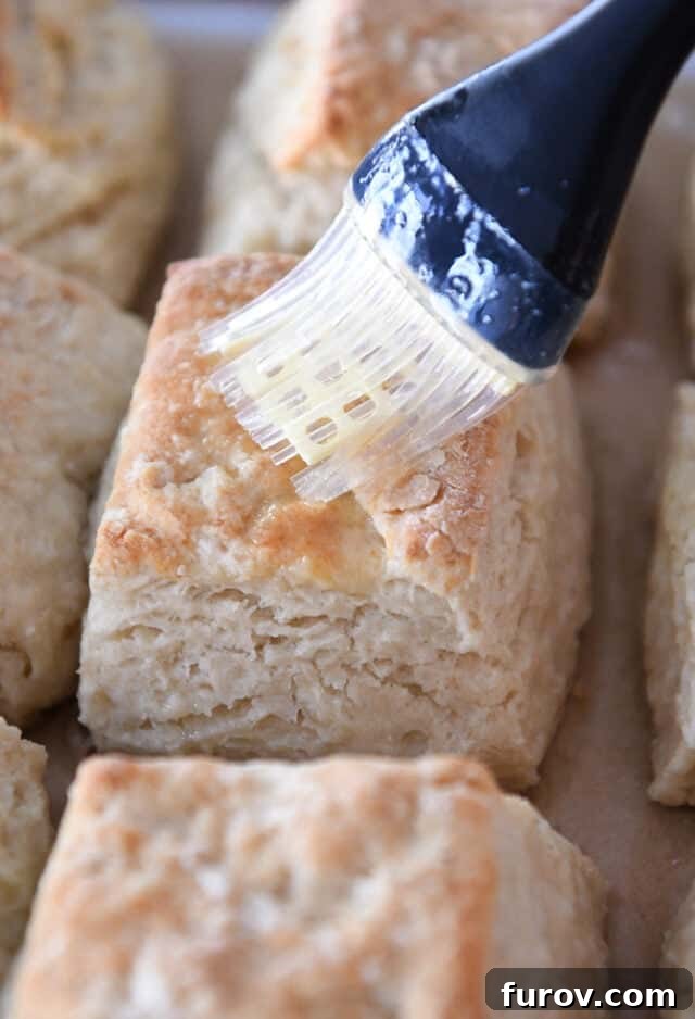 A close-up shot of a hand brushing melted butter onto the warm, golden tops of freshly baked sourdough biscuits, enhancing their flavor and appearance.