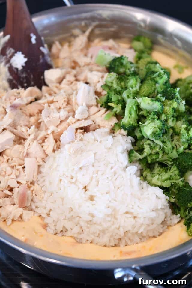 Cooked chicken, broccoli, and rice mixed into the rich cheese sauce in a large skillet, ready for transfer to a baking dish.