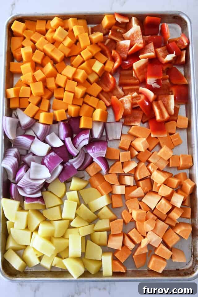 A baking sheet laden with uncooked butternut squash, sweet potatoes, red bell peppers, red onion, and Yukon gold potatoes, perfectly prepped and ready for roasting.