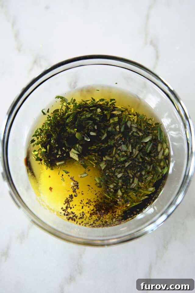 A glass bowl containing olive oil, balsamic vinegar, coarse salt, and sprigs of fresh thyme and rosemary, highlighting the key ingredients for the flavorful dressing.