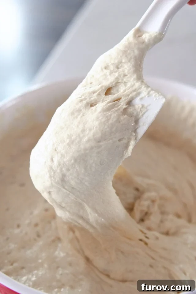 Rubber spatula stirring a bubbly, active sourdough starter in a glass jar.
