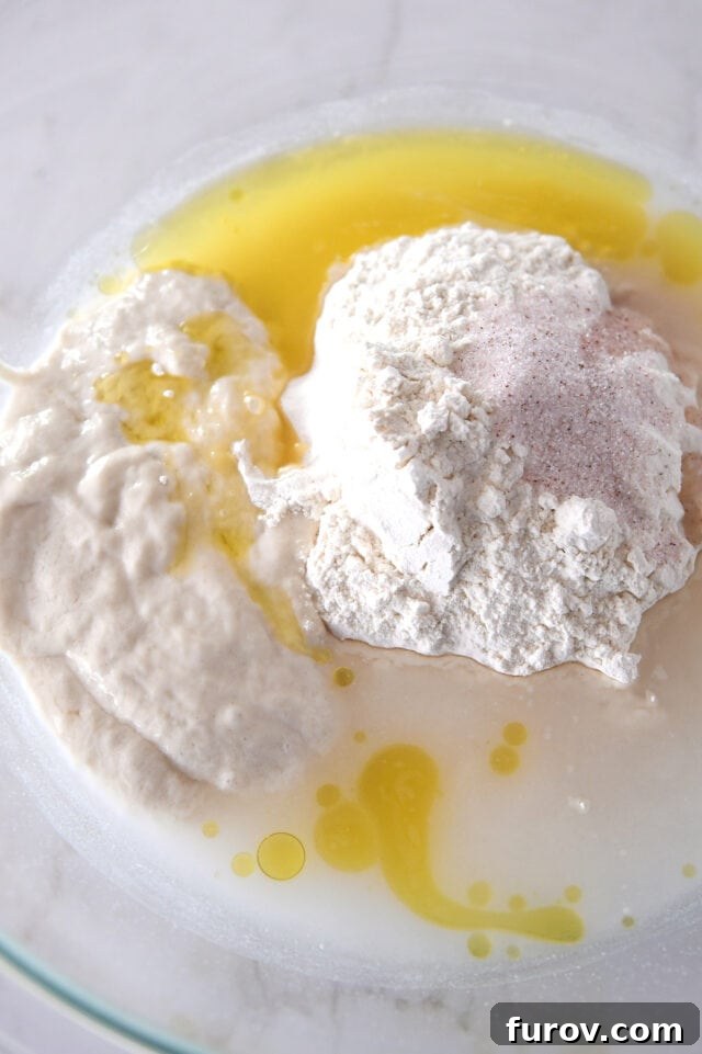 Glass bowl containing sourdough starter, flour, salt, olive oil, and water, ready to be mixed.