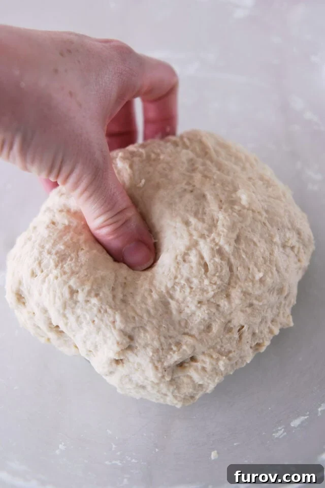 A thumb pressing into a soft ball of flatbread dough in a glass bowl, demonstrating ideal consistency.