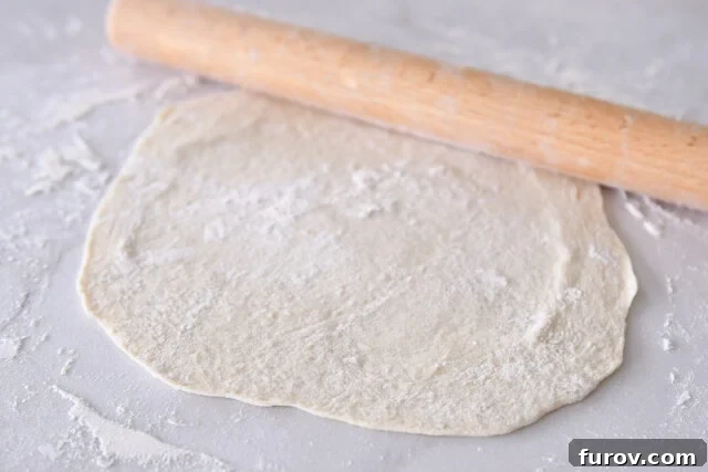Hands using a rolling pin to flatten sourdough flatbread dough into a thin circle on a floured counter.