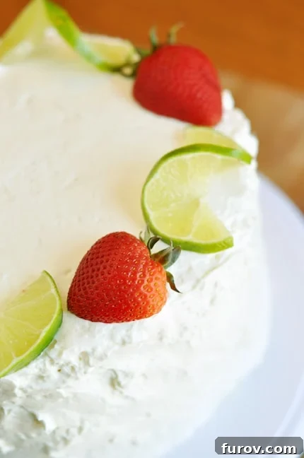 A beautifully frosted white cake on a pristine white platter, adorned with fresh strawberries and delicate ribbons of lime zest. The cake stands tall and inviting, promising a refreshing dessert experience.