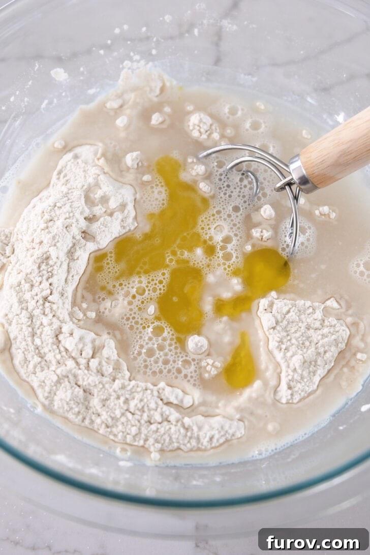 Dry and wet ingredients for bread dough in a glass bowl, with a Danish dough hook partially submerged.