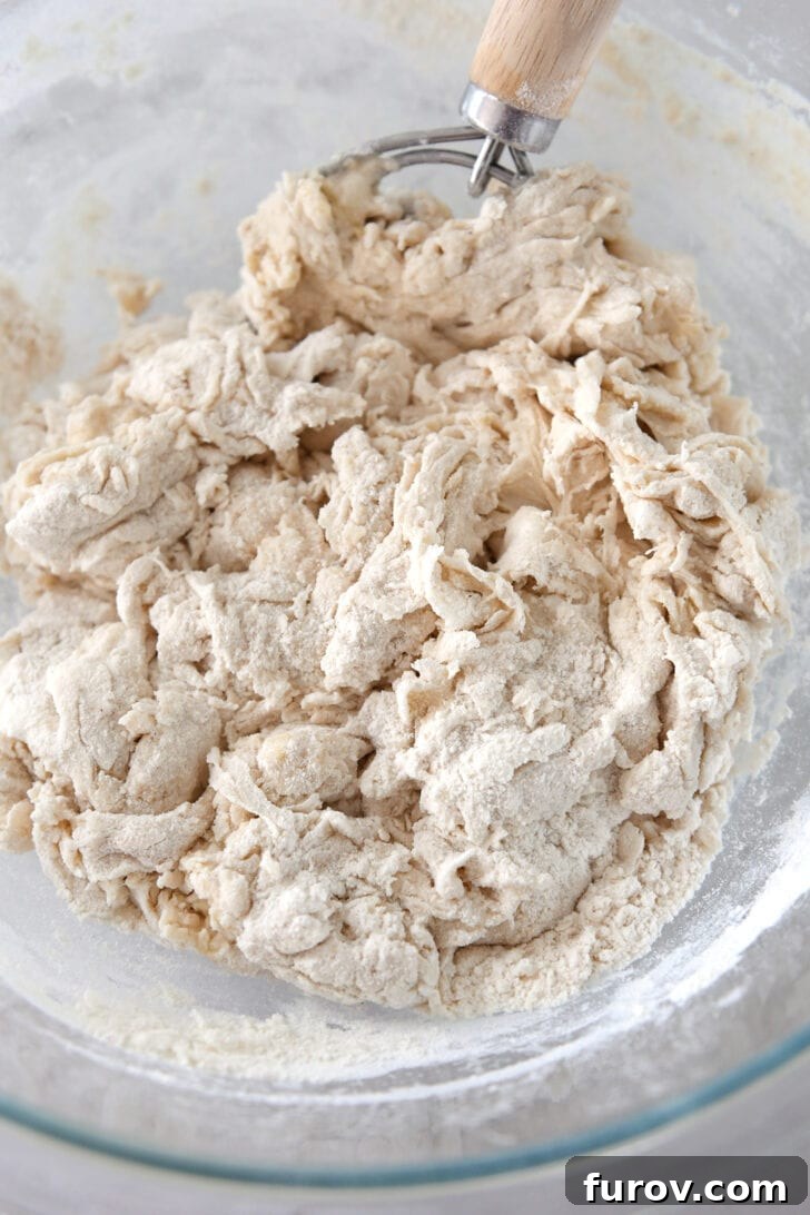 Bread dough ingredients partially mixed in a glass bowl with a Danish dough hook, showing the shaggy consistency.
