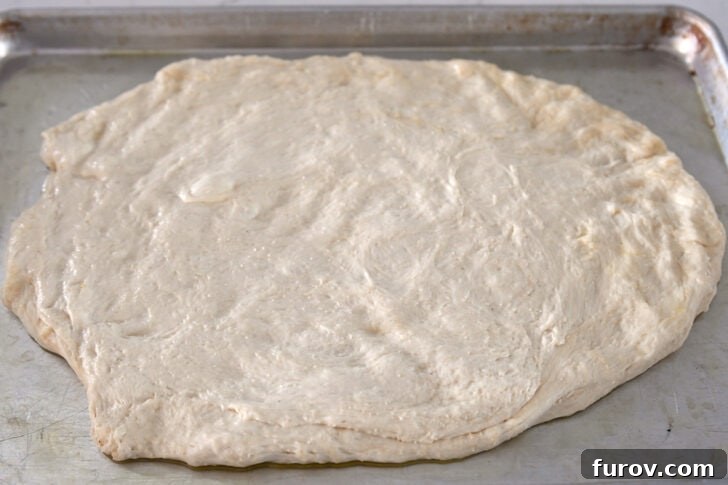 Bread dough gently pressed into a rough oval shape on a lightly greased half sheet pan, ready for toppings.