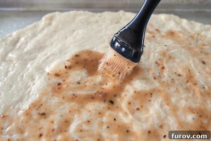 A pastry brush applying Italian salad dressing evenly over the surface of the shaped bread dough on a sheet pan.