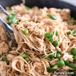 Close-up of kitchen tongs lifting a generous scoop of spicy garlic ramen noodles, rich with ground turkey, vibrant peas, and fresh green onions, from a skillet.