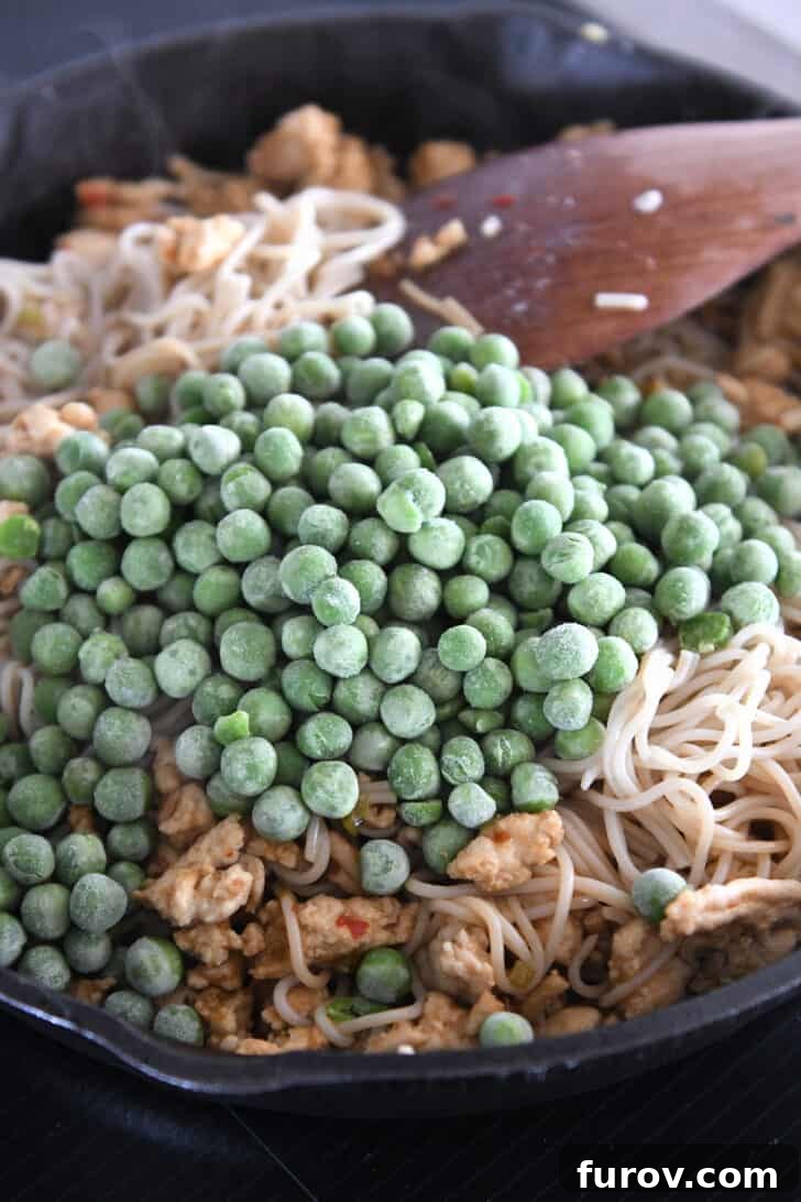 Frozen peas being added to a cast iron skillet, already filled with ramen noodles and ground turkey, introducing a fresh pop of color and sweetness.