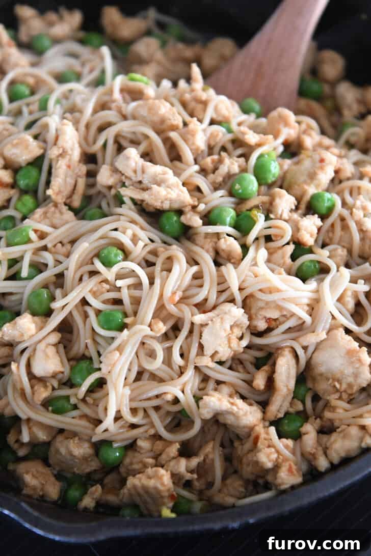 A pair of tongs expertly stirring ramen noodles, ground turkey, and frozen peas in a cast iron skillet, ensuring every ingredient is coated in the spicy garlic sauce.