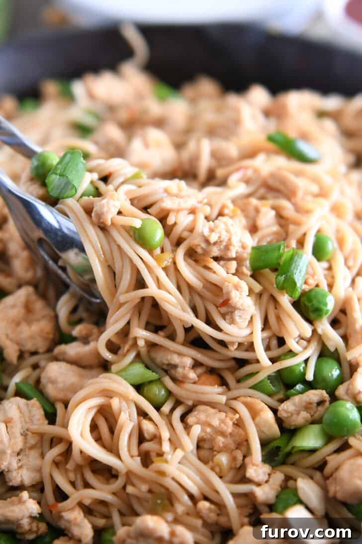 A close-up of kitchen tongs lifting a generous scoop of spicy garlic ramen noodles, rich with ground turkey, vibrant peas, and fresh green onions, from a skillet.