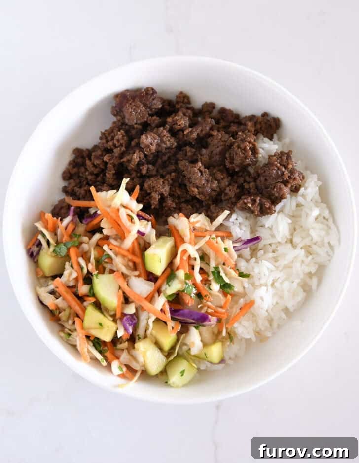 White bowl featuring rice, vibrant slaw, fresh cucumbers, and Korean ground beef, building a delicious meal.