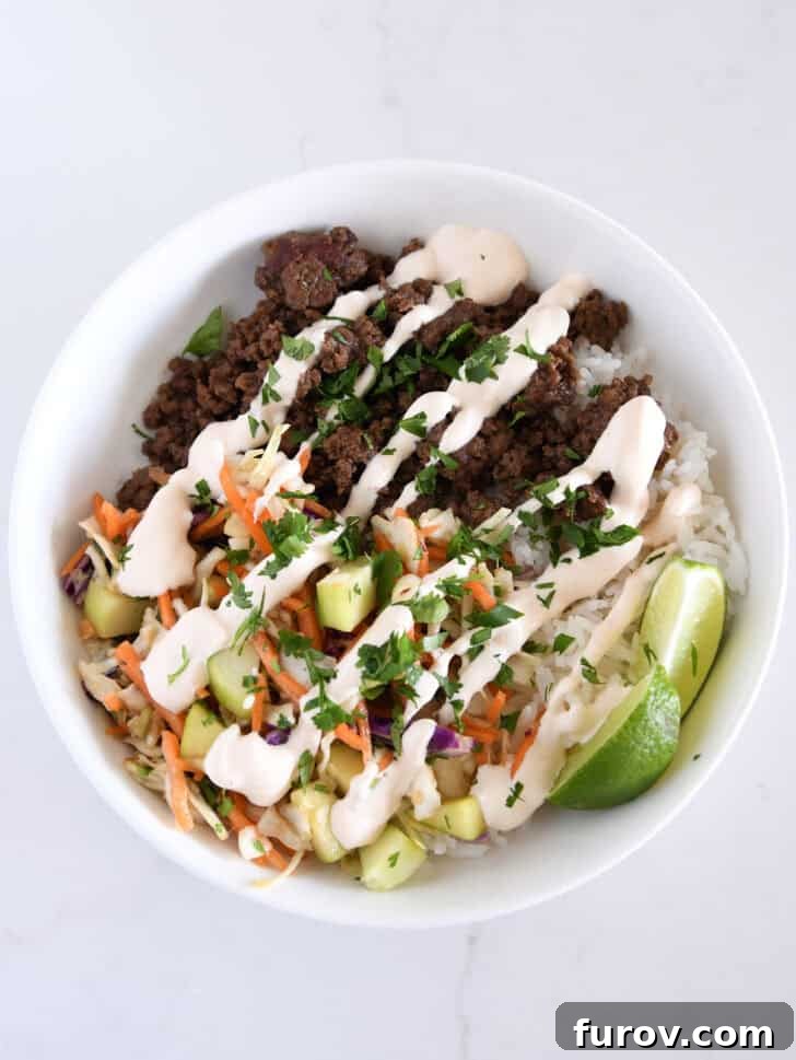 White bowl with rice, juicy lime wedges, fresh slaw, crisp cucumbers, flavorful Korean beef, and a drizzle of the creamy sauce.