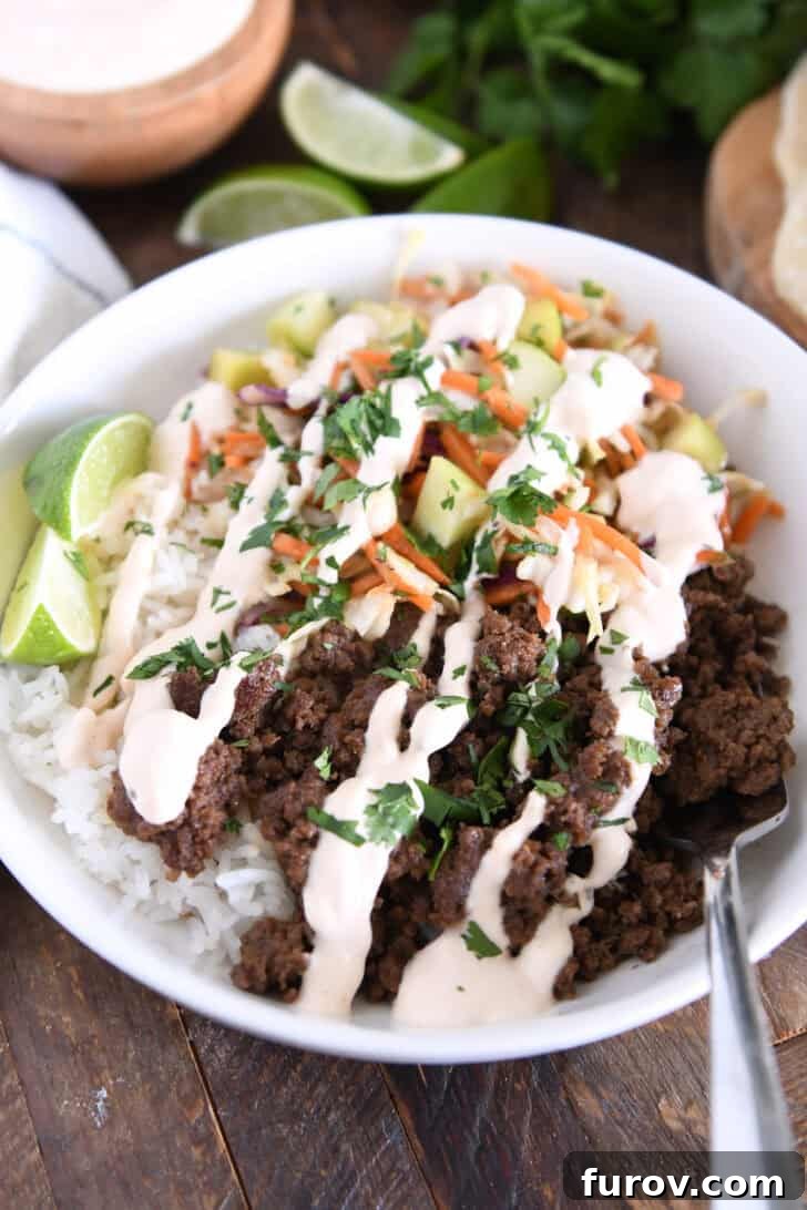 Fork in white bowl with rice, Korean beef, slaw, cucumbers, lime wedges and drizzled sauce, ready for a delicious bite.