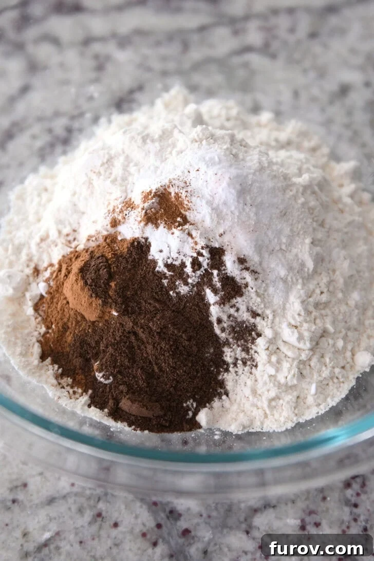 A clear glass bowl filled with all-purpose flour, ground cinnamon, baking powder, baking soda, and salt, representing the meticulously combined dry ingredients.