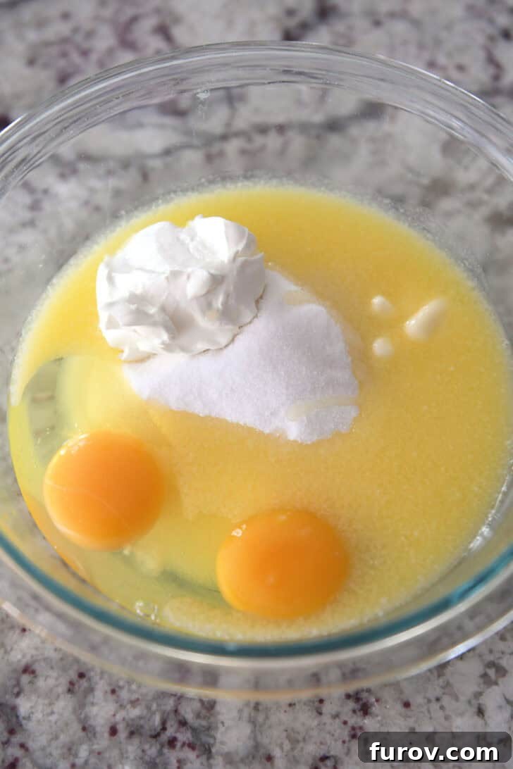 A transparent glass bowl containing whisked eggs, granulated sugar, creamy sour cream, and melted butter, representing the perfectly blended wet ingredients.