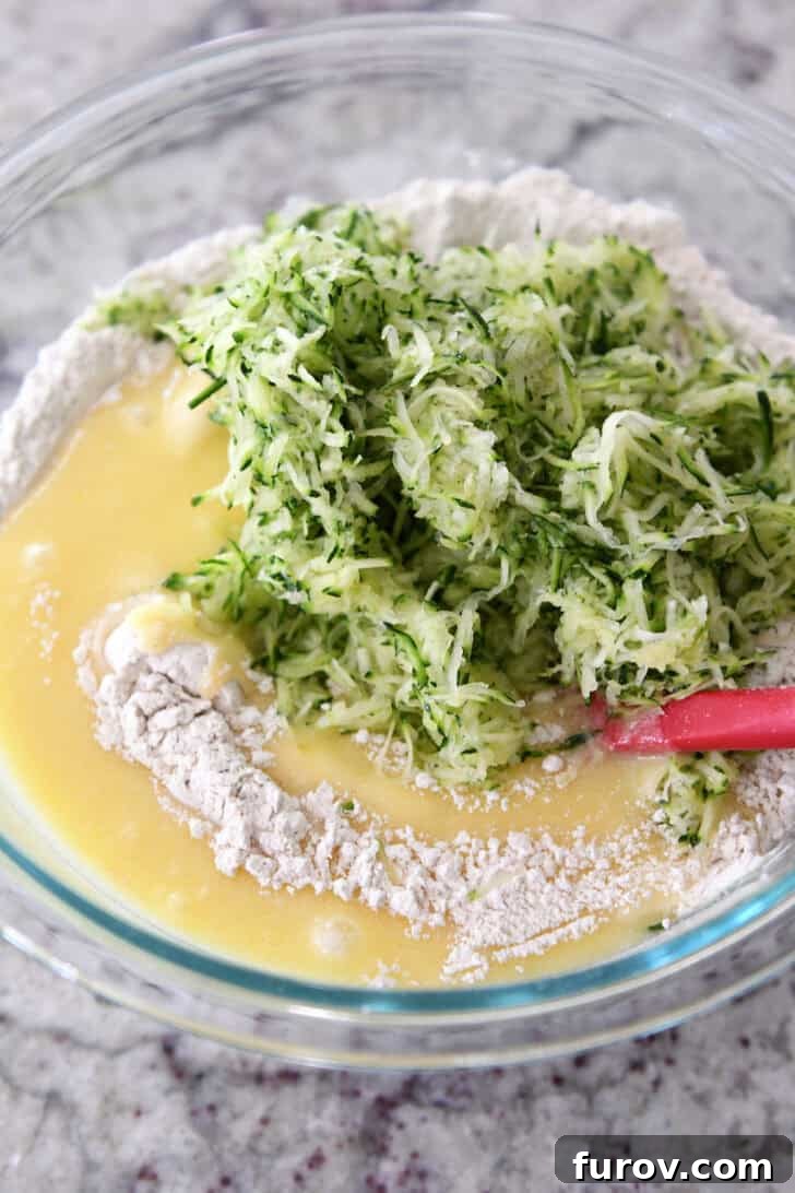 A large glass bowl holding the flour mixture, with the egg mixture and shredded zucchini added on top, illustrating the readiness for gentle folding.